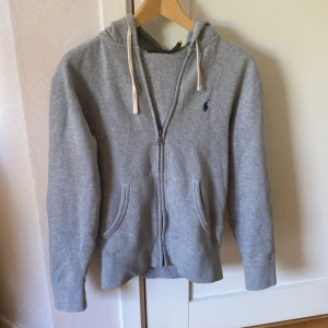 Grå hoodie från Polo Ralph Lauren - Säljer en klassisk grå hoodie från Polo Ralph Lauren med dragkedja och två fickor framtill. Tröjan har huva med vita snören och den ikoniska lilla loggan broderad på bröstet. Perfekt för en avslappnad stil.