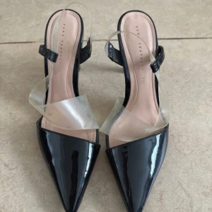 Svarta klackskor med transparent rem från Zara Trafaluc - Säljer ett par svarta klackskor från Zara Trafaluc i storlek 39. Skorna har spetsig tå, hög klack och en bred transparent rem över foten samt en smalare rem vid hälen. Snygg och modern design som sticker ut.