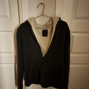 Mörkgrå hoodie med teddyfoder från Blend - Mysig mörkgrå hoodie från Blend med dragkedja och vitt teddyfoder på insidan. Jackan har huva och långa ärmar, perfekt för kyliga dagar. Enkel och stilren design som passar till det mesta. Pris kan diskuteras.