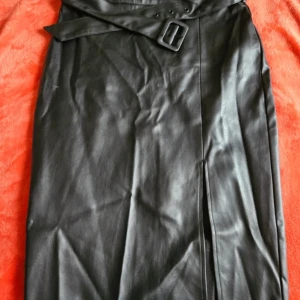 Svart skinnkjol med bälte och slits - Säljer en svart skinnkjol med snyggt bälte i midjan och en stilren slits framtill.  Och dom jag har aldrig använd bara finns inte prislapp.