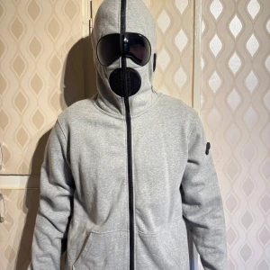 Grå hoodiejacka med inbyggda glasögon och mask - Unik grå hoodiejacka från Ai riders med hel dragkedja, inbyggda svarta glasögon och mask i huvan. Tröjan är till som skidtröja och är aldrig använd. Ny kostar 5 000