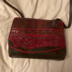 Brun och röd axelväska i skinn - Säljer en unik axelväska i brunt och rött skinn med dekorativa detaljer på framsidan. Väskan har flera fack med dragkedja både inuti och på baksidan, samt justerbar axelrem. Perfekt för dig som vill ha något annorlunda och snyggt till din outfit.
