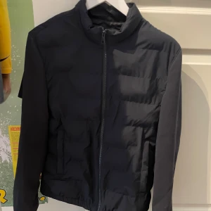 Svart pufferjacka med dragkedja - Svart pufferjacka med hög krage och hel dragkedja framtill. Jackan har två sidofickor och quiltad design som ger en sportig look. Perfekt för kyligare dagar och enkel att matcha med olika outfits.