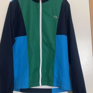 Färgglad vindjacka från Lacoste - Snygg vindjacka från Lacoste med cool färgblock-design i blått, grönt och mörkblått. Använd ett tag men är i jätte bra skick förutom där det står storlek syns även på bilden! Perfekt för dig som gillar sportig och stilren look. Pris kan diskuteras!!