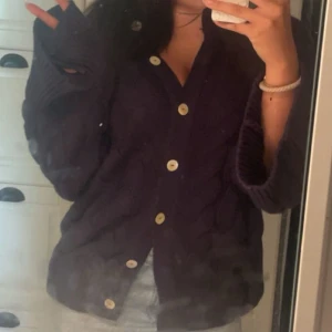 Mörkblå stickad kofta med knappar - Säljer en mysig mörklila stickad kofta med grova kabelstickade detaljer och stora ljusa knappar framtill. Koftan har långärmade, lite oversized ärmar och djup v-ringning. Perfekt att slänga på sig när det är kyligt ute. Är en L men passar mig och brukar ha s xs