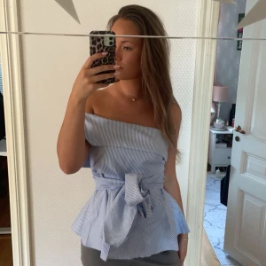 Blå och vit randig omlott topp - Säljer en snygg offshoulder topp med blå och vita ränder. Toppen har en bred knytning i midjan och peplum-formad nederdel som ger en cool siluett. Perfekt för dig som gillar att sticka ut med stilrena detaljer och offshoulder-look.