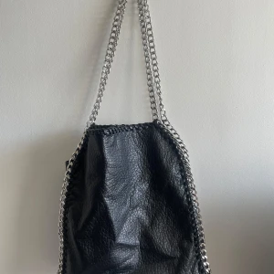 Svart axelväska med kedja från TIAMO - Cool svart axelväska från TIAMO i skinnimitation med silverfärgade kedjor som axelremmar. Nästan inget slitage förutom på en dragkedja som har lite rost