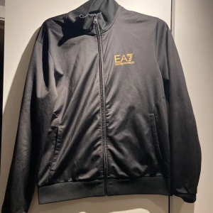 Svart EA7 jacka med guldlogga - Svart jacka från EA7 Emporio Armani med guldig logga på bröstet. Jackan har hel dragkedja, hög krage och två sidofickor. Tillverkad i ett mjukt och lätt material som känns sportigt och stilrent. Perfekt för dig som gillar clean och exklusiv streetstyle.