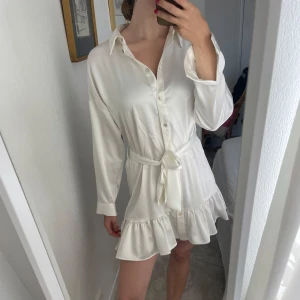 Vit kort skjortklänning med volang - Säljer en vit kort skjortklänning med volangkant och knytband i midjan. Klänningen har långa ärmar, klassisk krage och knappar framtill. Materialet är mjukt och har en lätt glansig finish som ger en lyxig känsla. Perfekt för dig som gillar stilrena och feminina plagg.