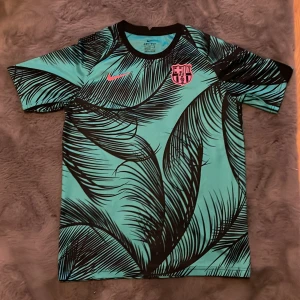Barca tröja med palm tryck  - Barcelona Dri-Fit fotbollströja från Nike i XL, med turkos bas och svarta palmbladsmönster. Rosa Nike-logga och Barça-emblem på bröstet. Rund hals och korta ärmar, tillverkad i lätt polyester som andas. Perfekt för dig som vill sticka ut på planen.