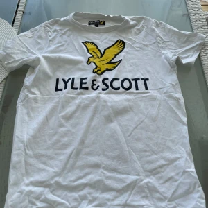 Vit Lyle & Scott t-shirt med tryck - Fräsch vit t-shirt från Lyle & Scott med stort tryck av gul fågel och svart logga på bröstet. Klassisk rund hals och korta ärmar. Perfekt för dig som gillar stilrena och sportiga märkesplagg. Mjuk och skön bomull som passar till jeans eller shorts.