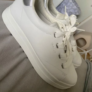 Vita sneakers från GANT med silverdetalj - Snygga vita sneakers från GANT med stilren design och platt sula. Skorna har breda vita snören och metallöglor för extra edge. Bakkappan har en cool silverdetalj som ger lite extra bling. Perfekta för dig som gillar cleana och fräscha sneakers i skinn.