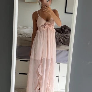 Ljusrosa långklänning - ljusrosa långklänning från Asos med märket Miss Selfridge med tunna justerbara axelband och v-ringad front. Klänningen har jättesöta blommor vid bysten och volangdetaljer längs kjolen. Tillverkad i ett tunt, flowy material som ger en drömmig känsla.