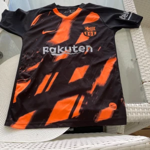 FC Barcelona Messi #10 svart/orange tröja - Snygg FC Barcelona fotbollströja från Nike med Messi #10 på ryggen. Svart bas med orangea grafiska detaljer och tryck, samt klubbmärke och sponsorloggor. Kortärmad modell i lätt och ventilerande material, perfekt för match eller träning.