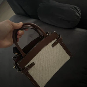 Beige och brun handväska från H&M - Snygg handväska från H&M i beige canvas med bruna detaljer och handtag i skinnimitation. Väskan har stilrena sömmar och en klassisk, boxig form som passar perfekt till din vardagsstil. Praktisk storlek för det viktigaste och enkel att matcha med olika outfits.