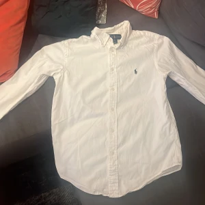 Vit skjorta från Ralph Lauren - Vit klassisk skjorta från Ralph Lauren med button-down krage och broderad logga på bröstet. Skjortan har lång ärm och knappar hela vägen framtill. Perfekt för dig som gillar stilrena och tidlösa plagg.