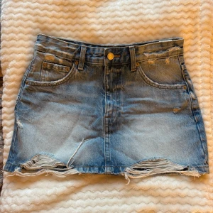 Blå jeanskjol från Pull&Bear - Snygg blå jeanskjol från Pull&Bear i storlek S. Kjolen har slitna detaljer och fransad nederkant för en cool vibe. Klassisk femficksmodell med knapp och dragkedja framtill. Perfekt för dig som gillar streetstyle och denim.