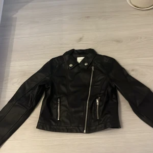 Svart skinnjacka från Lindex Kids - Snygg svart skinnjacka från Lindex Kids i storlek 140. Jackan har coola detaljer som sned dragkedja, två zip-fickor och klassisk krage med tryckknappar. Perfekt för dig som vill ha en edgy look och gillar streetstyle.