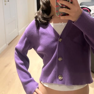 Lila croppad kofta med knappar - Superfin lila kofta med croppad passform och stora, glansiga knappar framtill. Tröjan har v-ringad hals och långa ärmar, perfekt att slänga över en topp. Mjuk stickad känsla och snygg vågig kant nedtill som ger extra stilpoäng. Använd en gång!