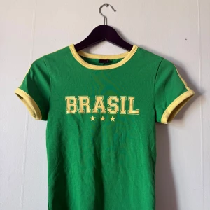 Grön Brasil t-shirt med gula detaljer - Snygg grön t-shirt med gul text 'BRASIL'. T-shirten har gula kantband vid hals och ärm. Hann tyvärr aldrig komma till användning så den är helt ny