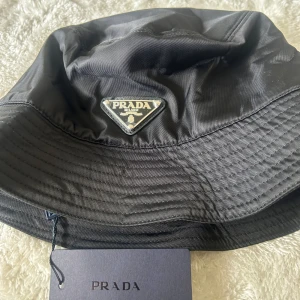 Svart bucket hat från Prada - Stilren svart bucket hat från Prada med ikonisk metallogo framtill. Hatten är tillverkad i polyester med bomullsfoder och har klassisk form med bred, nedåtlutande brätte. Perfekt accessoar för en trendig look.