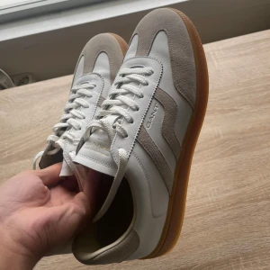 Vita och beige sneakers från Gant - Snygga sneakers från Gant i vitt skinn och beige mocka med klassisk gummisula. Skorna har rund tå, vita snören och stilrena detaljer som passar till många outfits. 
