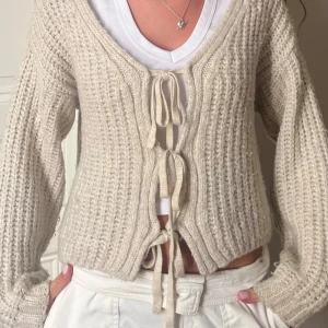 Beige stickad kofta med knyt - Mysig beige stickad kofta med v-ringning och tre knytband framtill. Koftan har lång ärm och en avslappnad. Köpt på Sellpy men är från asos 