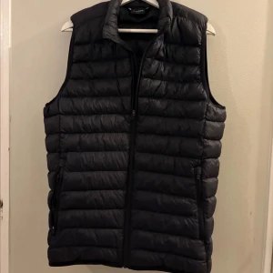 Svart dunväst med dragkedja - Säljer en svart dunväst från Everest med quiltad design och hög krage. Västen har två fickor med dragkedja och stängs med en hel dragkedja framtill. Perfekt att slänga över en hoodie eller tröja för extra värme.