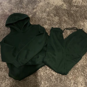 Mörkgrön hoodie med matchande byxor - Säljer en mörkgrön hoodie med huva och känguruficka, samt ett par matchande mjukisbyxor med snörning i midjan. Setet är gjort i mjukt och skönt material, perfekt för chill dagar eller när du vill vara extra bekväm. Passformen är normal och båda plaggen har en enkel, stilren look. Ny skick storlek S men passar de som har XS också 