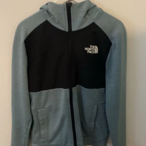 Blå och svart vindjacka från The North Face - Snygg hoodie med huva från The North Face i ljusblått och svart. Hoodien har dragkedja framtill, två fickor och logga på bröstet. Perfekt för dig som gillar streetstyle och vill ha en hoodie som funkar till många olika looks.