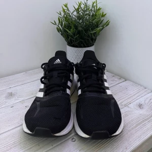 Svarta Adidas sneakers med vita ränder - Säljer ett par svarta Adidas sneakers med klassiska vita ränder på sidorna. Skorna har mesh-ovandel för bra ventilation, snörning och en platt sula med bra grepp. Perfekta för träning eller chill vardagsstil. Adidas-logga på plösen och snygg kontrast mellan svart och vitt.