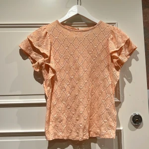 Persikofärgad blus Holly & Whyte XS - Supersöt persikofärgad blus från Holly & Whyte med korta volangärmar och ett snyggt quiltat mönster. Blusen har rund hals och knapp i nacken. Materialet är mjukt och strukturerat, perfekt för dig som gillar detaljer och en feminin vibe.
