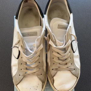 Philippe Model vita sneakers - Snygga vita sneakers från Philippe Model med grå mockadetaljer och svart häl. Klassisk rund tå och platt sula, med snörning framtill. Skorna har en unik skölddetalj på sidan och är tillverkade i skinn och mocka för en cool och urban vibe. Skorna har små hål på hälen av skon. Box ingår i köpet!