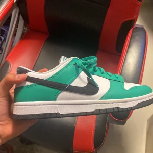 Nike Dunk Low vit och grön sneaker - Nike Dunk Low sneakers i fräsch vit och grön färg med svart swoosh på sidan. Skon har snörning, platt sula och rund tå. Ovandelen är i syntet och skinn, med klassisk Nike-logga på tungan och hälen. Perfekt för dig som gillar streetstyle och vill sticka ut.