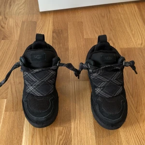 Svarta sneakers från UGG är endast använda en gång!!!!!!! - Säljer ett par svarta UGG sneakers med coolt rutmönster på ovansidan och breda snören. Skorna har en robust platt sula med UGG-logga undertill och mjukt foder på insidan. Materialmix av mocka och syntet ger både stil och komfort. Perfekt för dig som gillar streetwear.