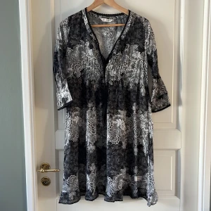 Odd Molly svartvit mönstrad klänning - Svartvit kortklänning från Odd Molly med blommigt och paisleyinspirerat mönster, broderad fågel på ryggen och dekorativ spetskant vid halsringningen. Klänningen har trekvartsärm och är tillverkad i tunn, luftig bomull. Perfekt för dig som gillar unika detaljer och boho-vibe.