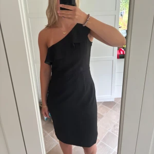 Svart oneshoulder klänning By Malina - Snygg svart klänning från By Malina med oneshoulder och volangdetalj över axeln. Modellen är knälång och figursydd. Storlek M, jag är storlek S i vanliga fall så denna är lite stor på mig. Helt ny med prislapp!