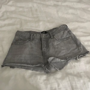 Grå jeansshorts från 157 - Snygga grå jeansshorts från 157 med råa, fransiga kanter och klassisk femficksdesign. Modellen har knappgylf och bälteshällor, perfekt för dig som gillar en chill och edgy stil. Passar dig som vill ha korta shorts med loose fit och cool vibe.