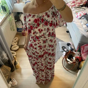 Vit långklänning med röda rosor - Superfin offshoulder långklänning i vit med röda rosor och gröna blad. Klänningen har en romantisk volang upptill, slits framtill och är axelbandslös. Perfekt för sommarens alla tillfällen! Materialet känns lätt och luftigt.