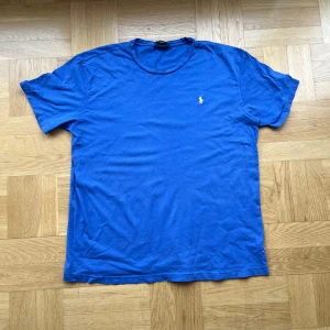 Blå t-shirt från Polo Ralph Lauren - Blå t-shirt från Polo Ralph Lauren med klassisk rund hals och korta ärmar. T-shirten har en liten broderad logga i vitt på bröstet och är gjord i mjuk bomull. Perfekt för dig som gillar enkel och stilren design. Obs har en  fläck längst ner till höger.