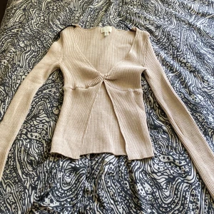 Beige ribbad omlottopp från H&M - Säljer en beige ribbad omlottopp från H&M i storlek S. Toppen har lång ärm, v-ringad hals och snygg twistad detalj framtill som skapar en öppen effekt under bysten. Materialet är mjukt och stretchigt