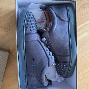 Mörkgrå Christian Louboutin sneakers - Säljer ett par mörkgrå sneakers från Christian Louboutin i mocka med coola nitar på tån och klassisk röd sula. Skorna har snörning och rund tå, samt en platt sula. Perfekt för dig som vill sticka ut med exklusiv stil och edgy detaljer.