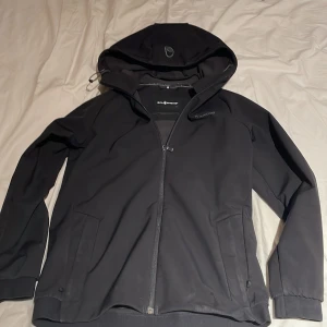 Svart vindjacka från Sail Racing - Svart vindjacka från Sail Racing med huva och hel dragkedja framtill. Jackan har diskreta loggor på bröstet och huvan, samt två sidofickor. Materialet är syntet och jackan har en stilren, sportig look som passar perfekt för blåsiga dagar. Skulle säga är som xs-s fast det står 176