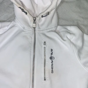 Sail racing tröja - Vit hoodie från Sail Racing med hel dragkedja framtill och en extra zipficka på bröstet. Snyggt broderad logga på framsidan och justerbar huva med snörning. Mjuk bomullskänsla och långärmad modell, perfekt för chill dagar.