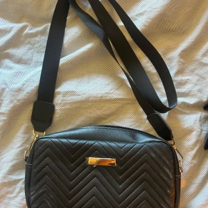 Svart axelväska med gulddetalj - Snygg svart axelväska med quiltat zigzag-mönster och en stilren guldfärgad detalj framtill. Väskan har en justerbar axelrem och dragkedja upptill. Perfekt storlek för att få plats med det viktigaste och passar till många olika outfits. Aldrig använd🩷