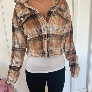 Rutig teddyjacka från SHEIN, S - Supermjuk och croppad teddyjacka från SHEIN i brun, beige och orange rutig design. Jackan har långa ärmar, stora bröstfickor med knappar och är gjord i fluffig polyester. Perfekt för dig som gillar en chill och trendig look.