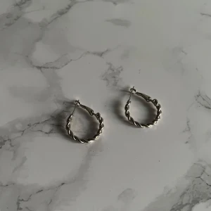 Silvriga tvinnade hoops örhängen - Säljer ett par snygga silvriga hoops med tvinnad design. Perfekta för dig som gillar stilrena smycken med lite edge. De är runda och har en enkel låsning som gör dem lätta att ta på och av. Passar till alla outfits och ger en cool touch.