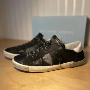 Philippe Model svarta sneakers strl 43 - Svarta sneakers från Philippe Model och coolt sköldmärke på sidan. Skorna har snörning,med fin text på samt platt sula och detaljer i både skinn och mocka. Snygg kontrast mellan svart och vitt, perfekt för dig som gillar stilrena och unika sneakers.