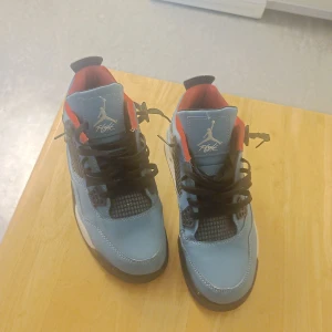 Nike Jordan 4 Retro University Blue - Säljer ett par Nike Jordan 4 Retro University Blue sneakers med ljusblå mocka, svarta och vita detaljer samt röd insida. Skorna har snörning, meshpaneler och speckled svart häl med röd Jumpman-logga. Perfekt för dig som vill sticka ut med en ikonisk modell.