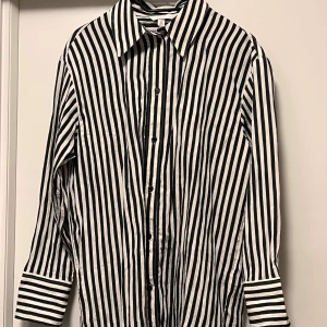Svartvit randig skjorta från H&M - Säljer en svartvit skjorta från H&M i storlek S. Skjortan har vertikala ränder, klassisk krage och knappar hela vägen framtill. Långärmad med snygga manschetter. Perfekt för dig som gillar en stilren och cool look.